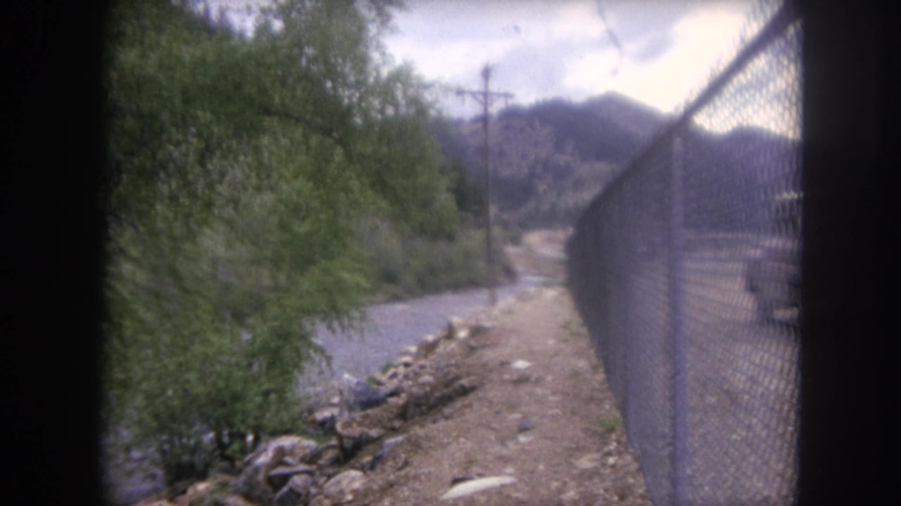 1962 Winter Park, Colorado travel footage frame with a chain-link fence, riverbank, trees, and mountain terrain.