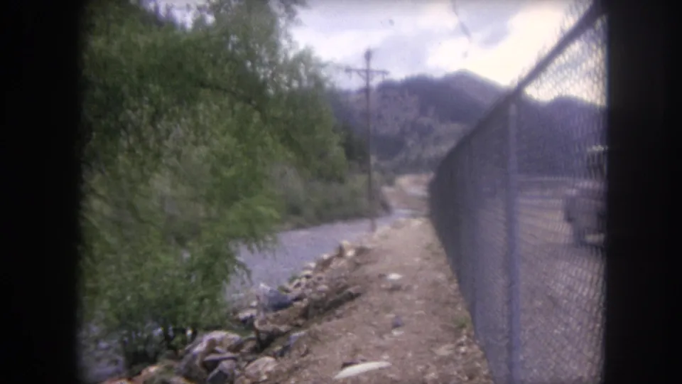 1962 Winter Park, Colorado travel footage frame with a chain-link fence, riverbank, trees, and mountain terrain.