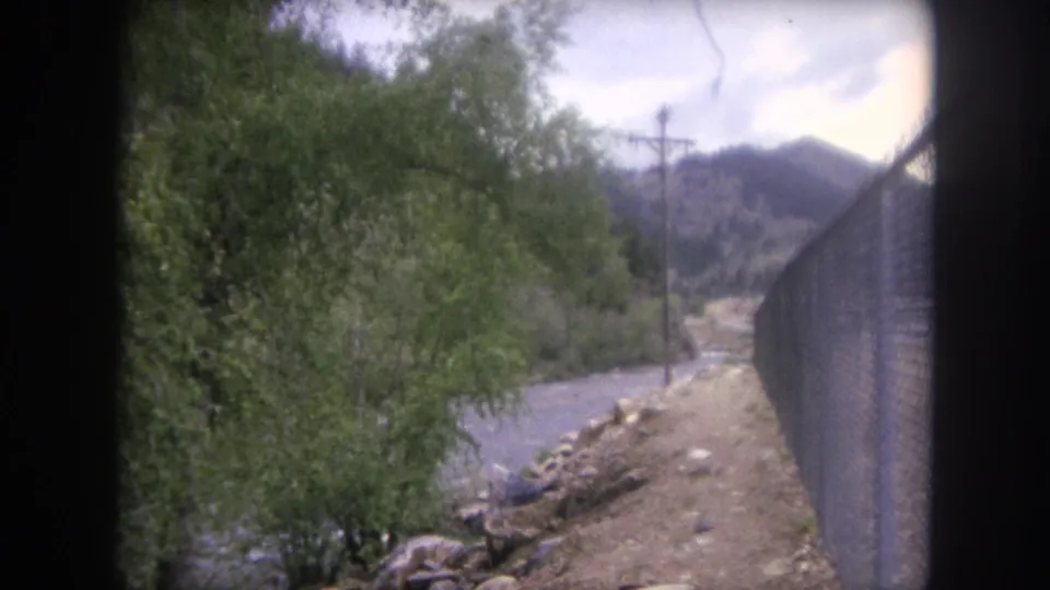 Winter Park, Colorado 1962 home-movie frame with trees, a river corridor, fence line, dirt shoulder, and mountain background.