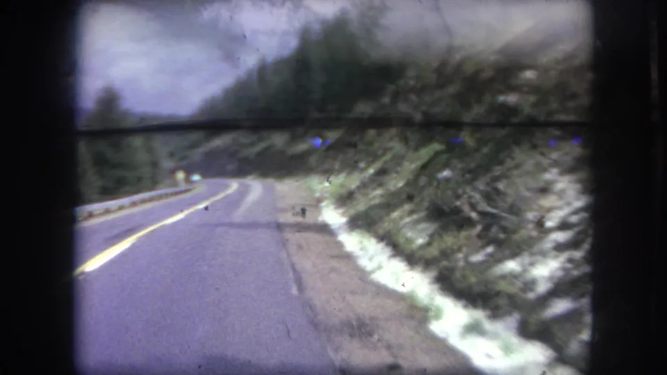 1962 Winter Park, Colorado driving view on a curving mountain road with yellow centerline, trees, and rocky hillside.