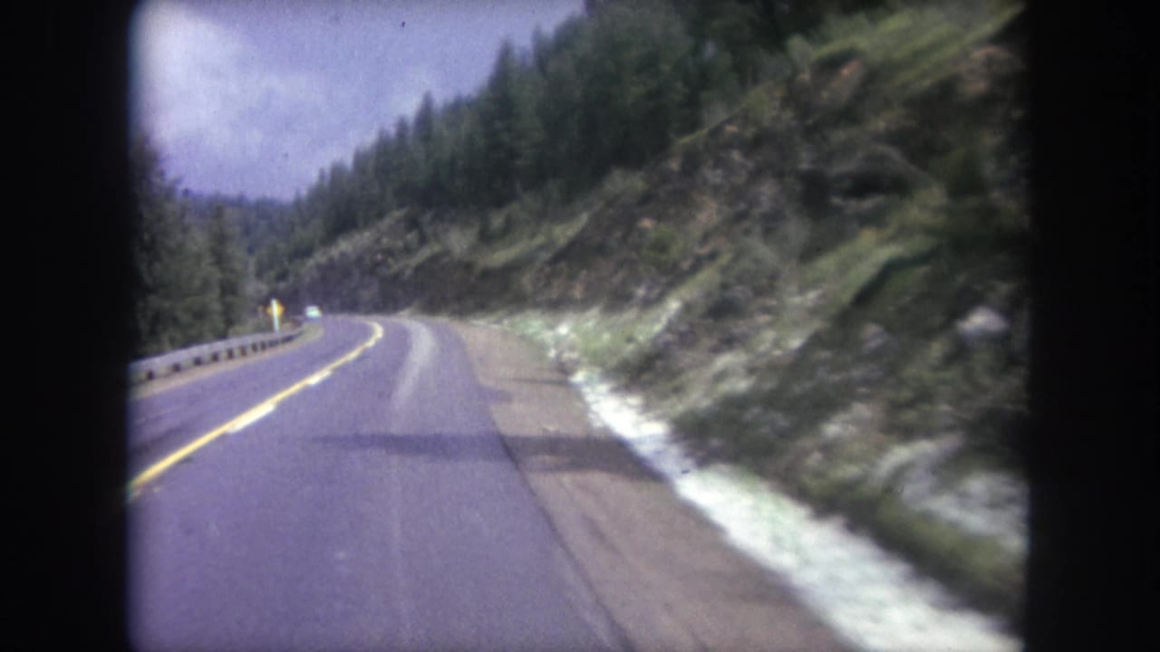 Winding two-lane mountain highway with yellow lines, guardrail, trees, and rocky slope.