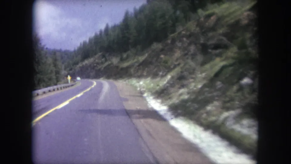 1962 Winter Park, Colorado home-movie view of a curving mountain highway bordered by guardrail, evergreens, and rocky roadside slope.