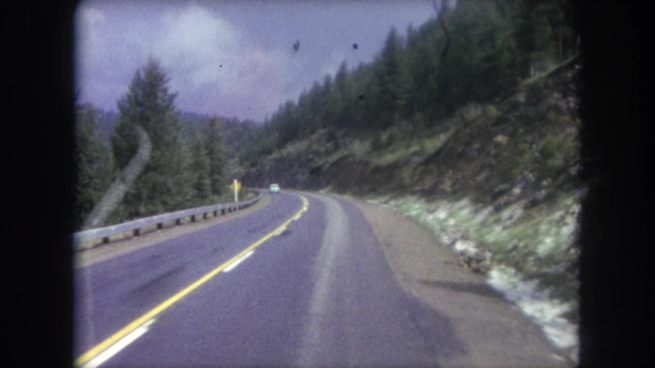 Forward-facing 8mm view along a curving Winter Park mountain highway, with a distant car, guardrail, forest, and rocky road cut.