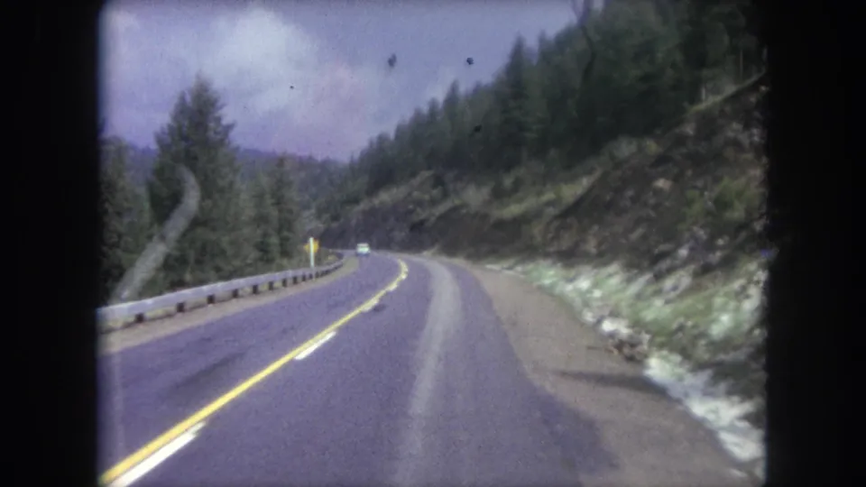 Forward-facing 8mm view along a curving Winter Park mountain highway, with a distant car, guardrail, forest, and rocky road cut.