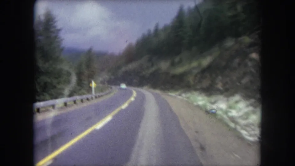 Soft 8mm frame from a moving car on a 1962 Colorado mountain road, showing a guardrail, curve, forested slope, and car ahead.