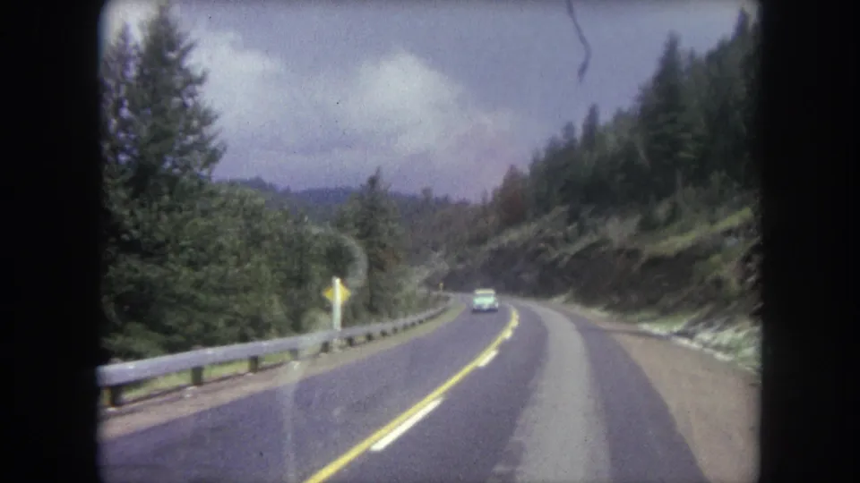 Winter Park mountain highway scene with a distant car, left-side guardrail, yellow roadside sign, forested slopes, and cloudy sky.