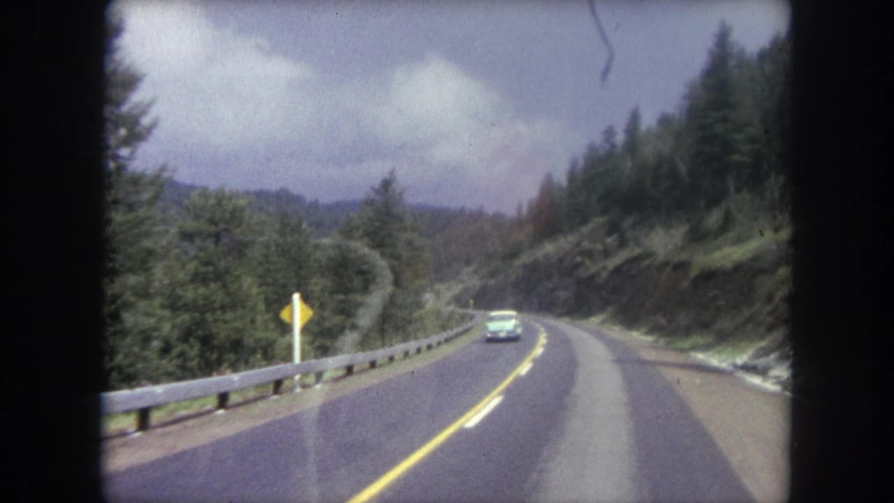 1962 8mm view from inside a vehicle on a winding mountain road near Winter Park, Colorado, with trees and an oncoming car ahead.