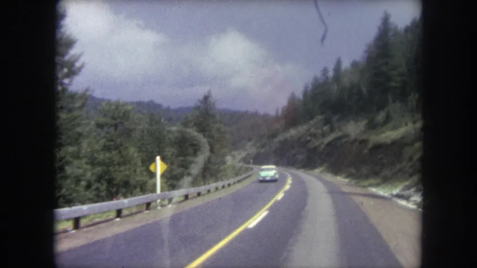 1962 8mm view from inside a vehicle on a winding mountain road near Winter Park, Colorado, with trees and an oncoming car ahead.