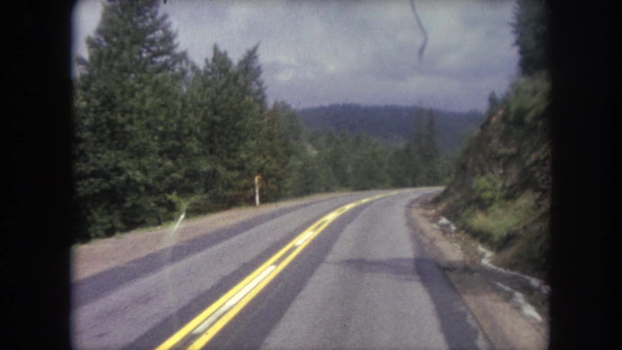 Passenger-view 8mm frame of a Winter Park, Colorado mountain road curving through dense evergreens under an overcast sky.