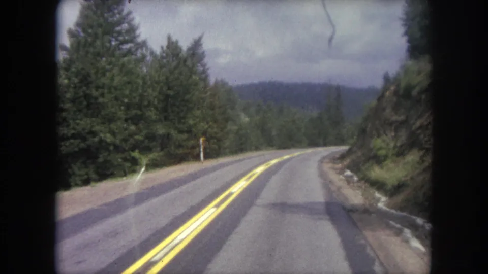 Passenger-view 8mm frame of a Winter Park, Colorado mountain road curving through dense evergreens under an overcast sky.