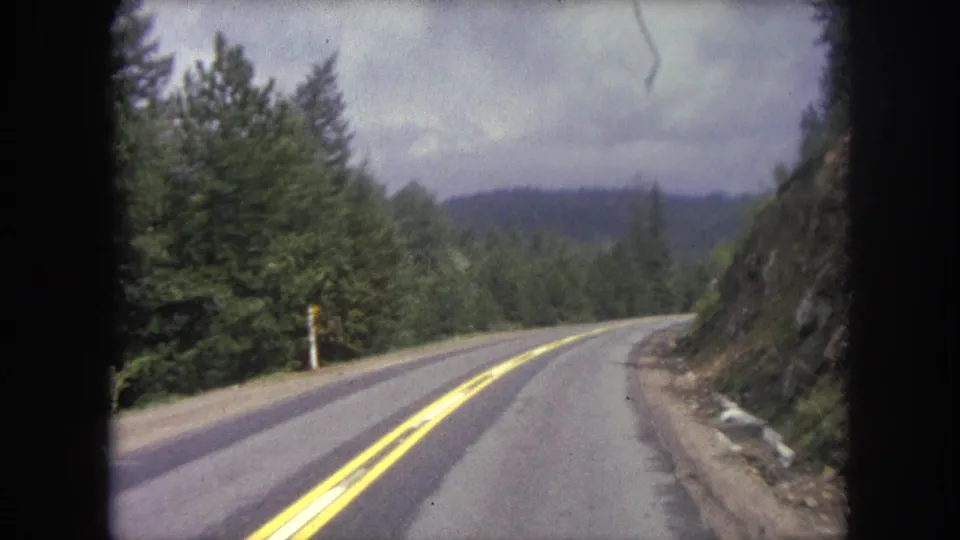 1962 home-movie frame from a moving car on a forested mountain road in Winter Park, Colorado, with road markings and hillside visible.