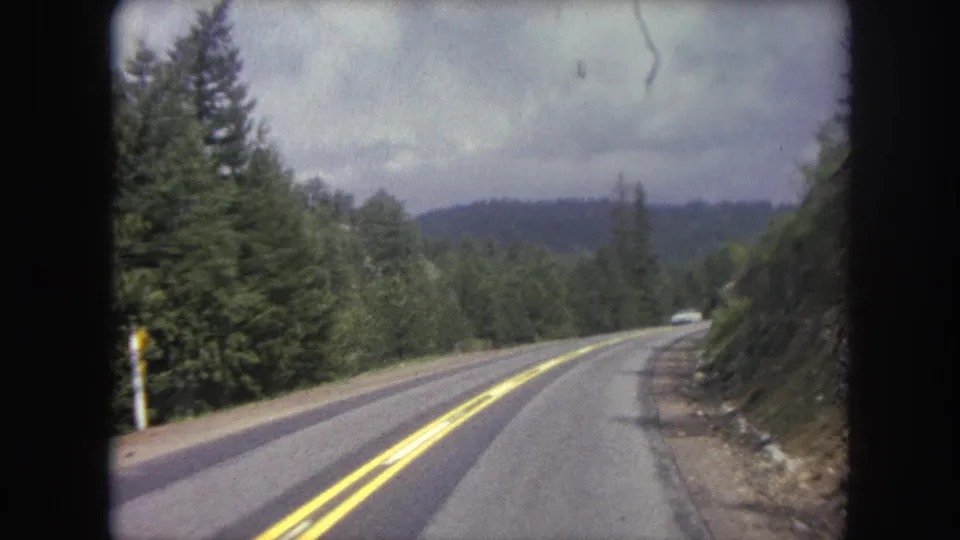 Final 8mm frame of the Winter Park mountain drive, showing a curving road, evergreen forest, and a small distant car near the bend.