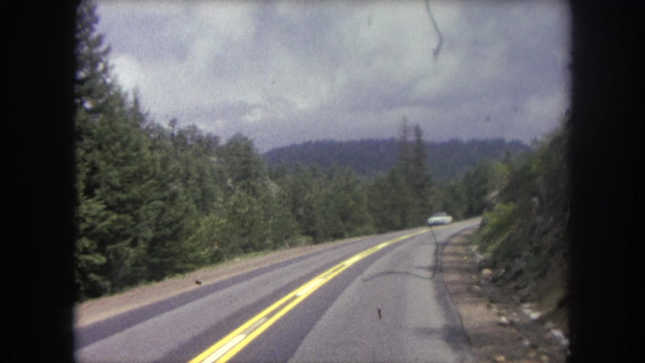 Two-lane mountain road seen through a car window, with forested hills and a distant vehicle.