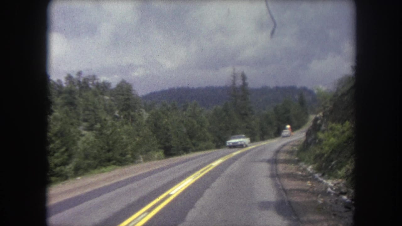 A classic car approaches along a two-lane road near Winter Park, Colorado, with forested hills and cloudy sky beyond.