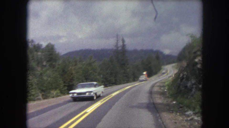 A white early 1960s-style sedan passes the moving camera on a curving road in the Winter Park, Colorado area.