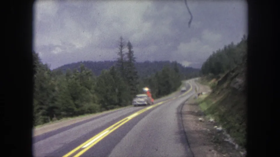 A car and a bright red roadside vehicle light appear along the two-lane Winter Park road, framed by forest and hills.
