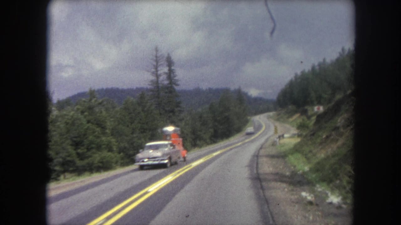 Two-lane mountain road through evergreen forest with an oncoming sedan towing an orange trailer.