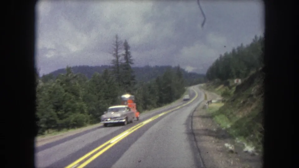 1962 Winter Park, Colorado home-movie view of a winding two-lane road with an oncoming sedan and bright orange trailer.