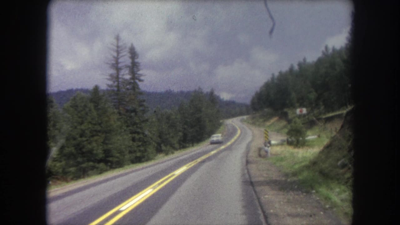 Moving-vehicle view in Winter Park, Colorado, showing a curving mountain road, forested slope, distant car, and roadside marker.