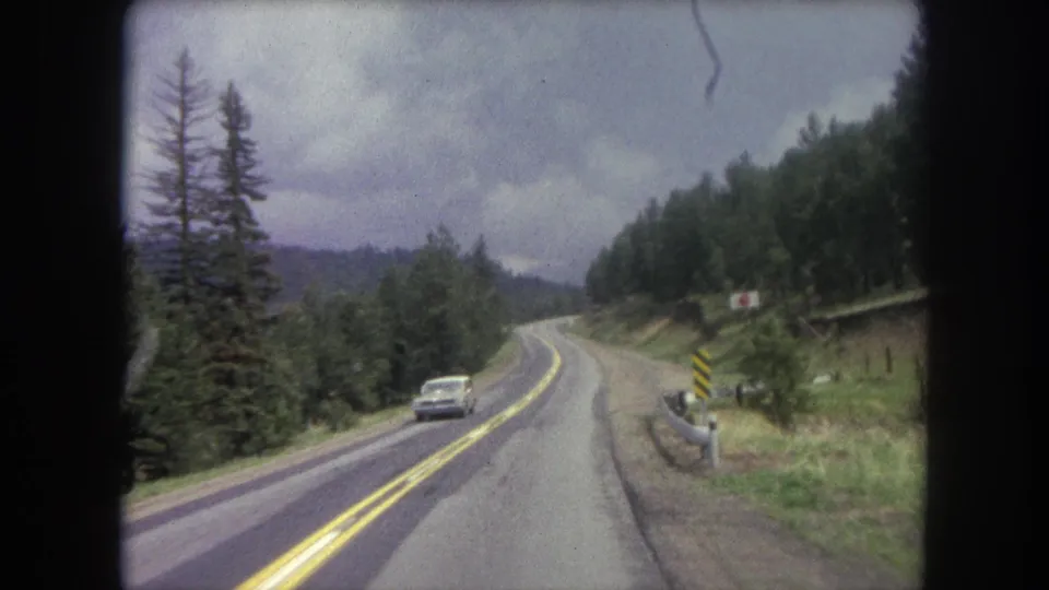 1962 Colorado road scene with a light-colored oncoming car, double yellow lines, trees, hillside, and striped shoulder sign.