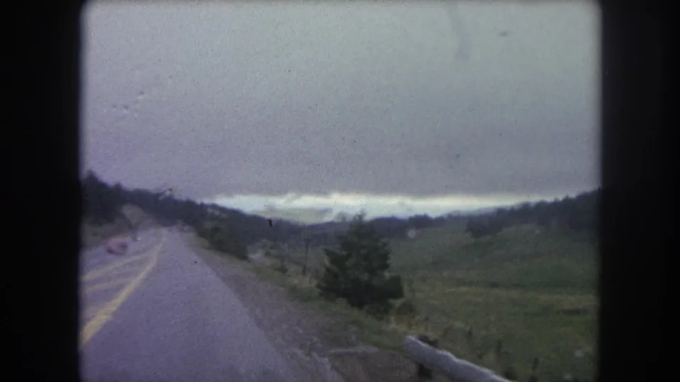Winter Park area moving-road view showing a gray sky, mountain ridges, open green field, roadside shoulder, and guardrail end.
