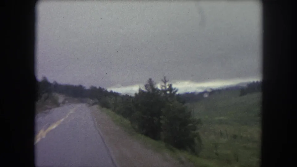 1962 home-movie road view near Winter Park, Colorado, showing a two-lane road, bushes, fields, and distant hills.