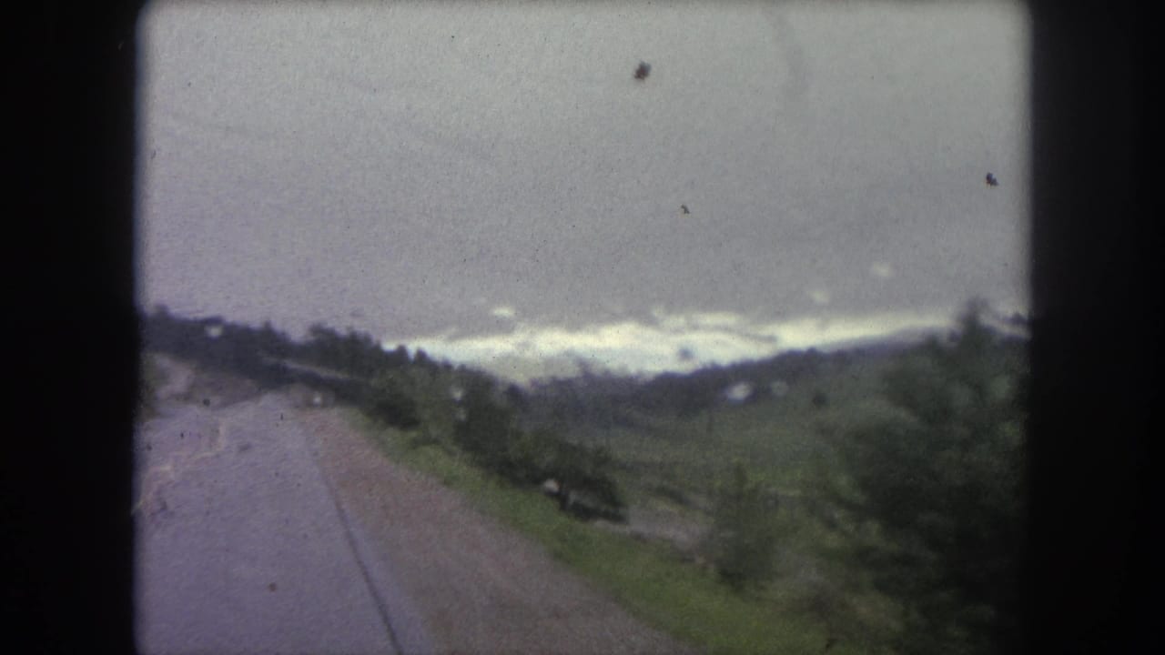 Moving-car view of a wet road and green mountain slope in Winter Park, Colorado, with a gray sky and soft 8mm color.