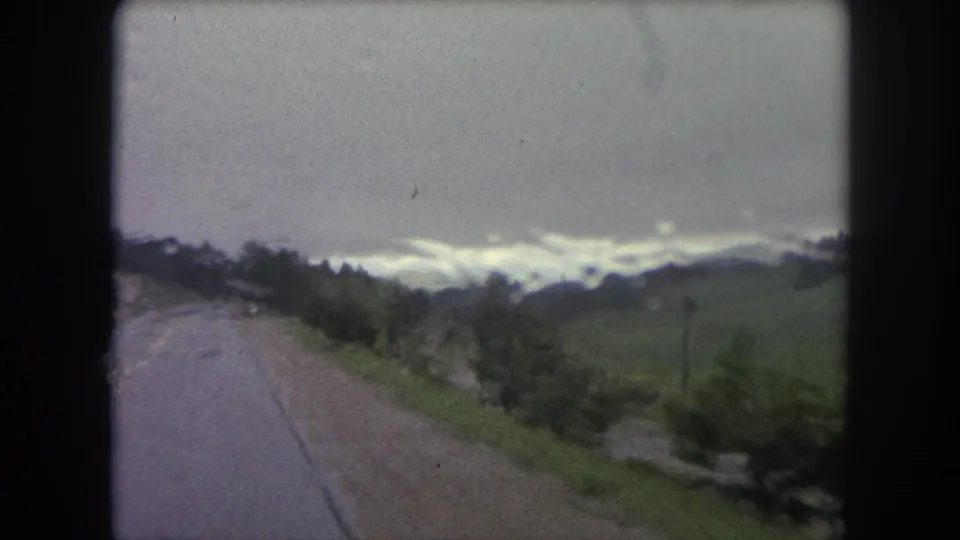 1962 home-movie frame looking along a wet Colorado road toward rolling green hills and a low, cloudy ridgeline.