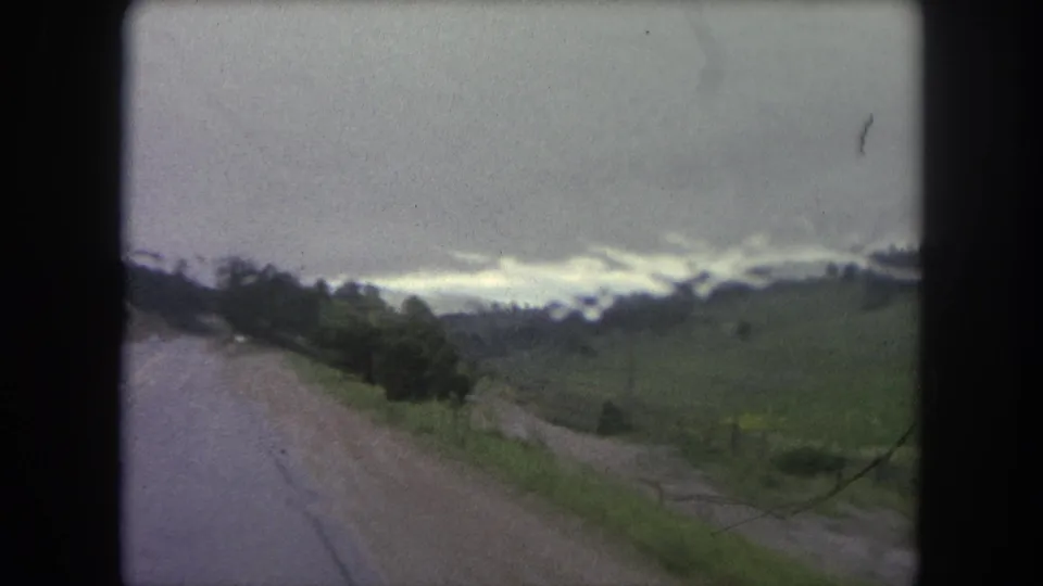 End frame of the 8mm Winter Park road view, with a wet roadside, open green hillside, scattered trees, and overcast mountains.
