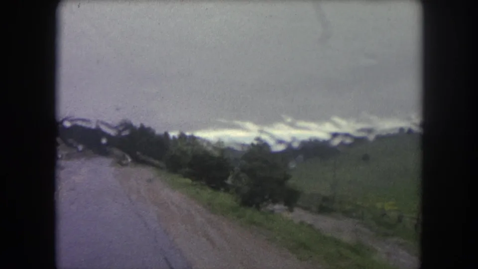 1962 Winter Park, Colorado home-movie frame showing a wet dirt road, roadside trees, green slopes, and cloudy sky.