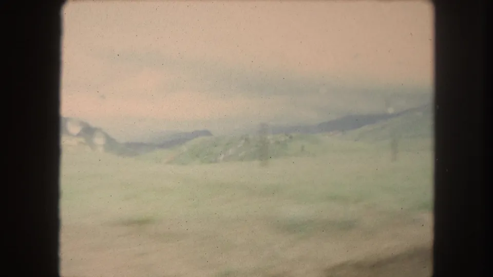 1962 Winter Park, Colorado frame with an open field, distant ridge, green hillside, and dark vehicle window frame at the side.
