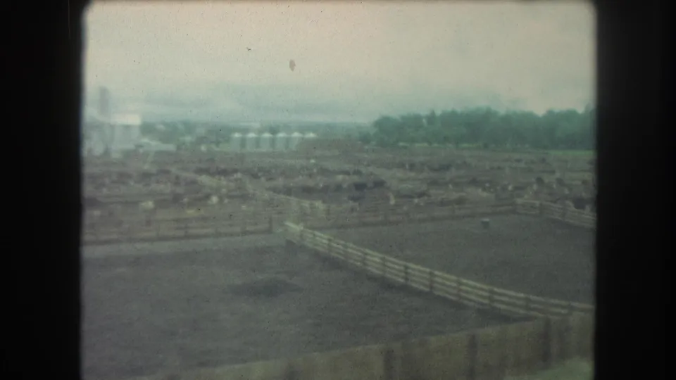 Aerial 8mm frame of the Winter Park ranch landscape, with wooden fences, livestock areas, distant trees, and pale overcast sky.