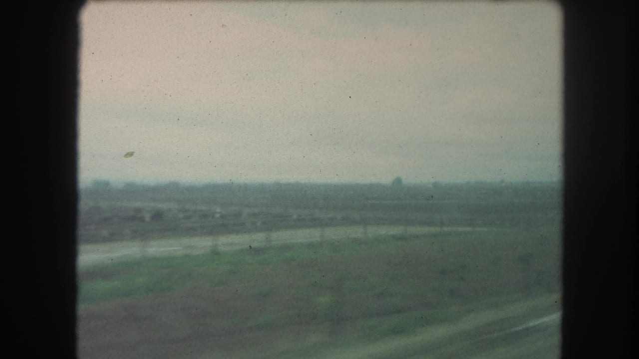 1962 8mm home-movie frame of flat farmland and a distant tree line, viewed from a moving vehicle in the stated Winter Park, Colorado area.