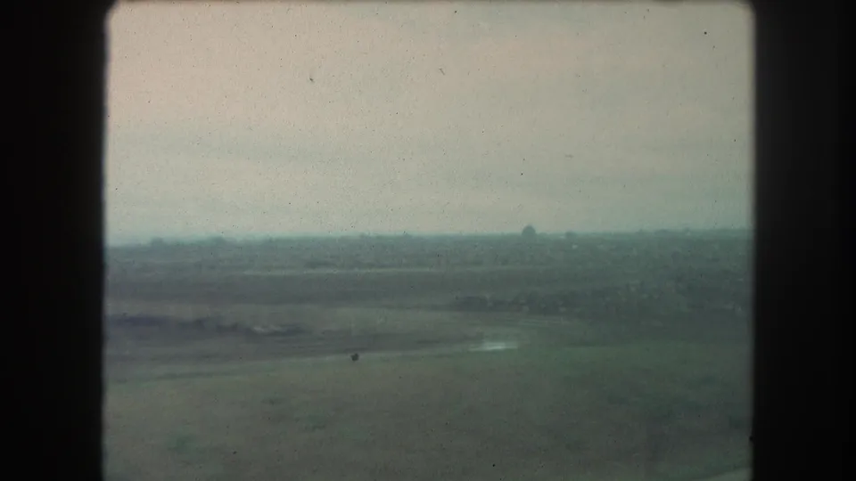 Grainy 8mm frame showing flat fields, a pale road or watercourse, and distant tree line from a moving travel viewpoint.