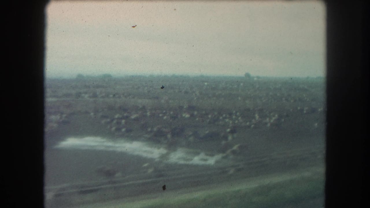 1962 Winter Park, Colorado train-window frame with open land, a reflective water area, transport lines, and small buildings in the distance.