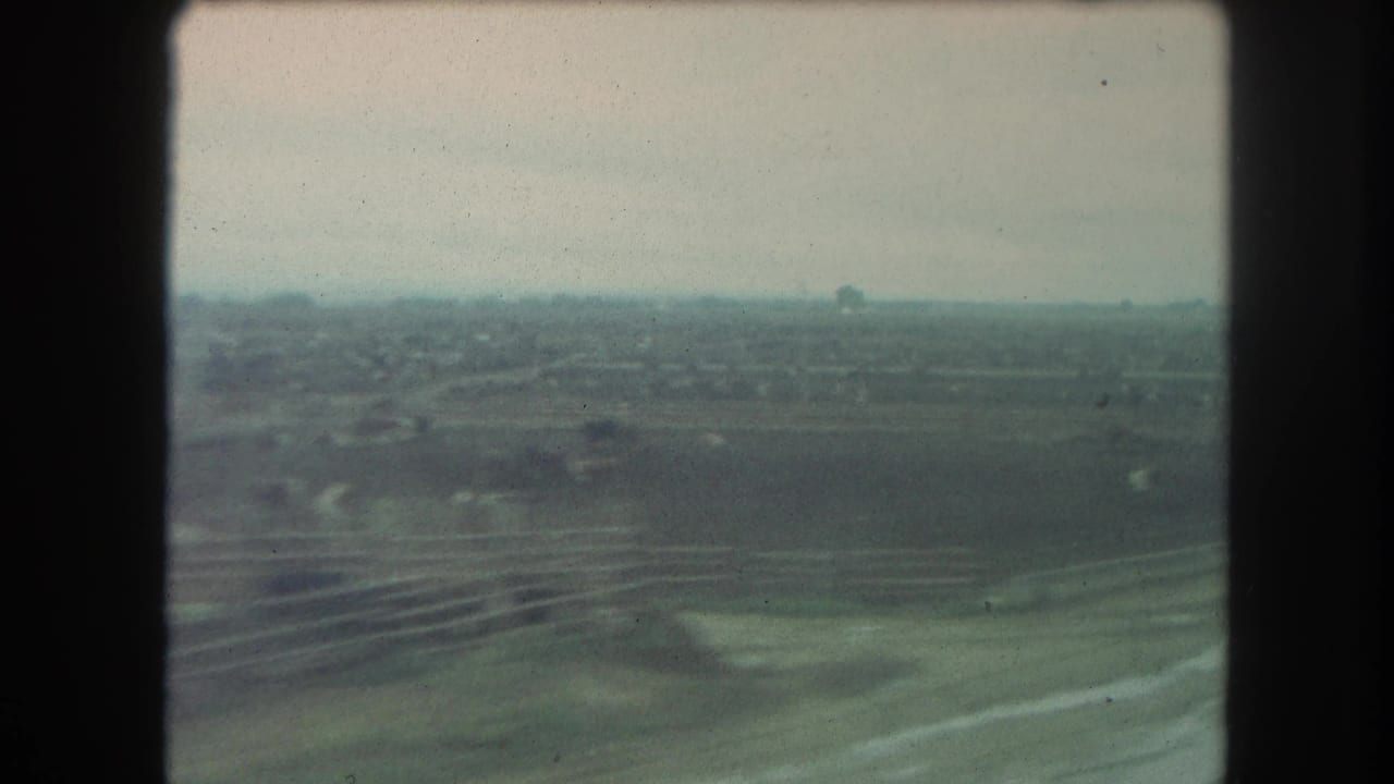 Train-window view over hazy rural fields with distant trees and scattered structures.