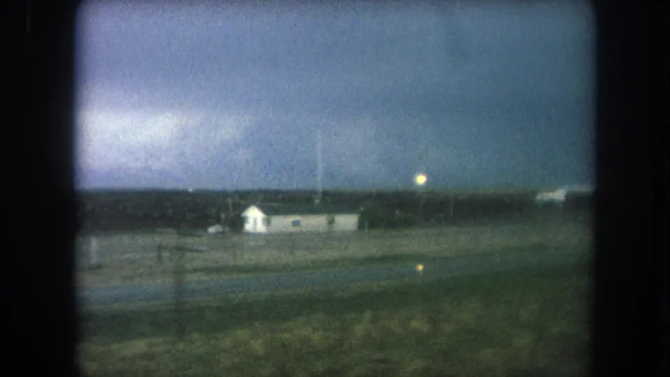 1962 home-movie frame showing a small white house near open countryside and roadside utility poles at dusk.