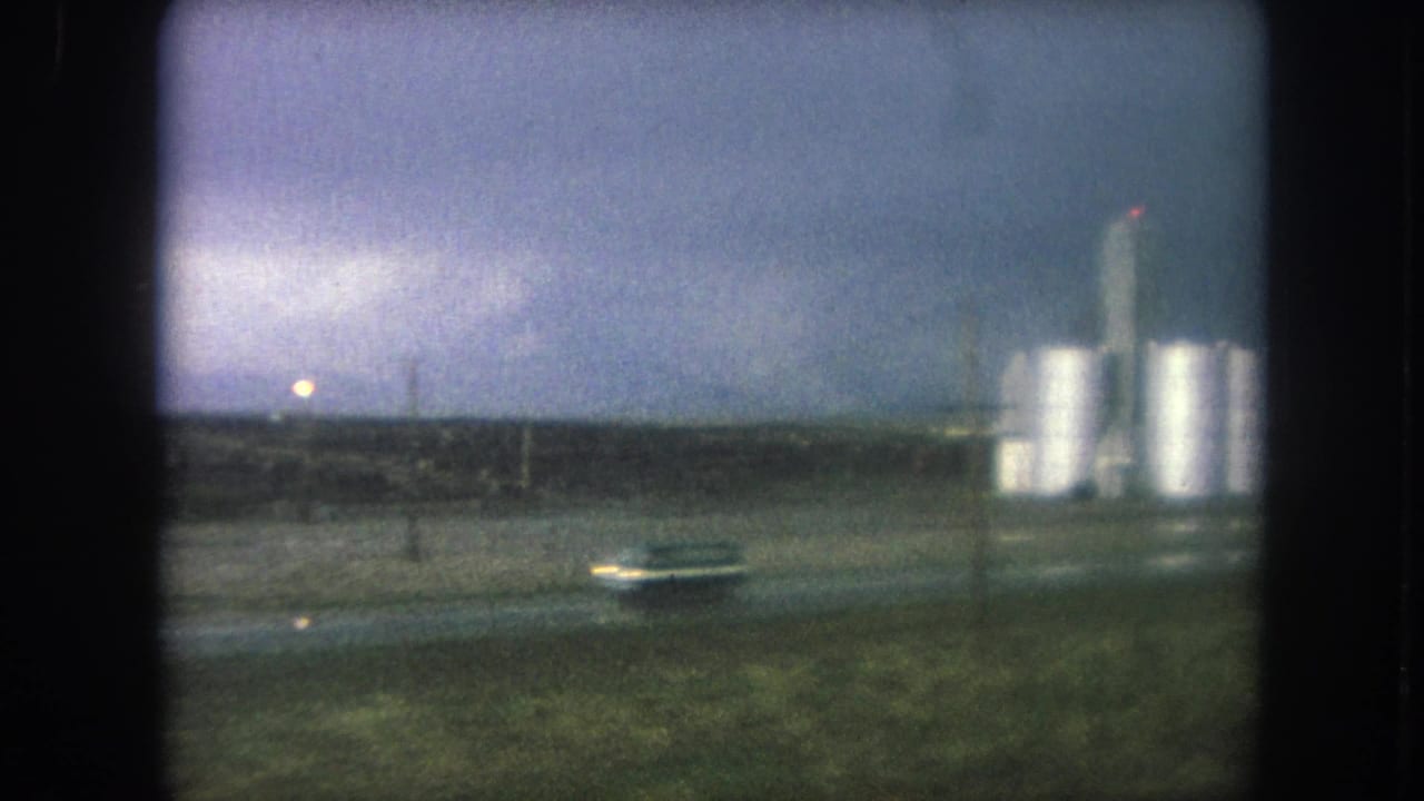 Distant industrial silos beside a wet roadside under a dark cloudy sky.