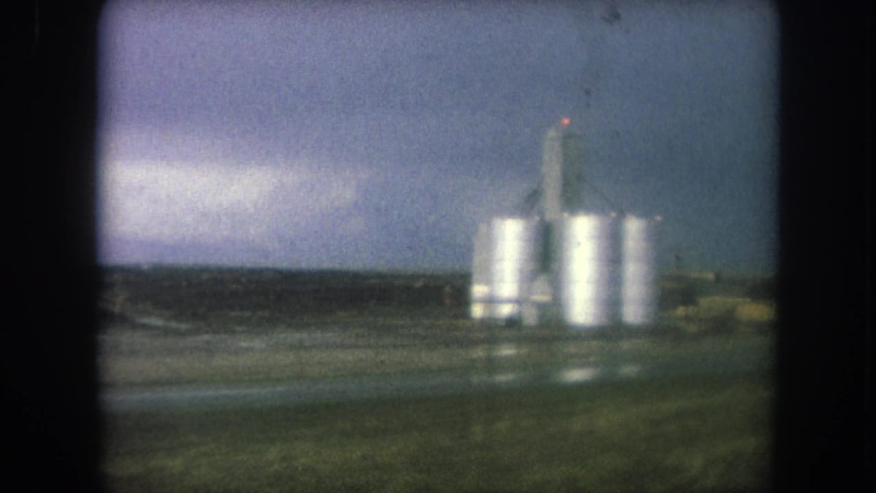 Cluster of silver silos and a tall tower seen across wet roadside land.
