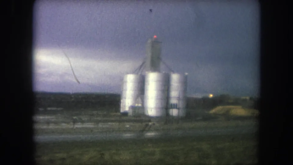 Closer 1962 8mm frame of industrial silos in Winter Park, with utility poles, reflective wet ground, and a gloomy sky.