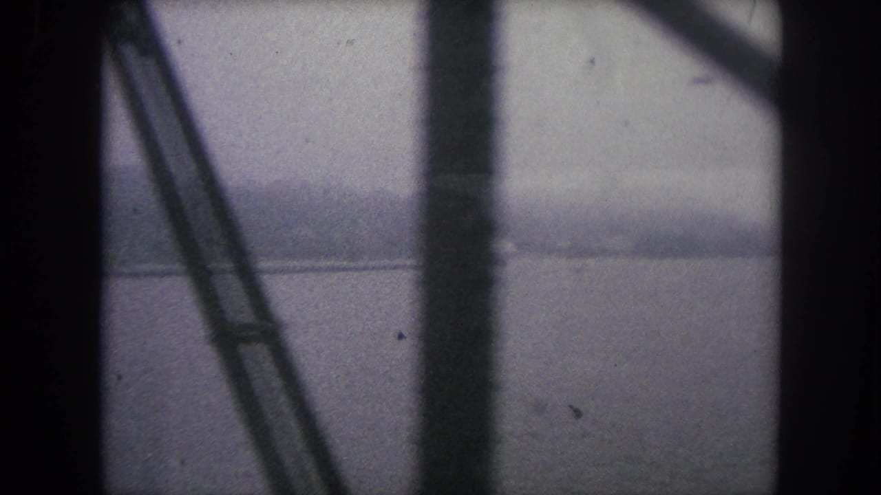 Muted 1962 8mm frame of water and hazy land partially obscured by dark vertical and diagonal supports.