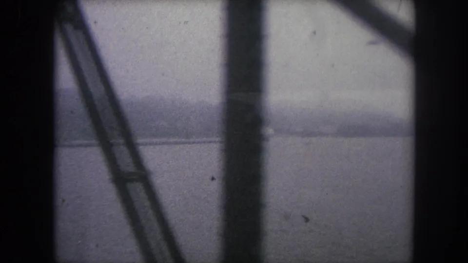 Muted 1962 8mm frame of water and hazy land partially obscured by dark vertical and diagonal supports.