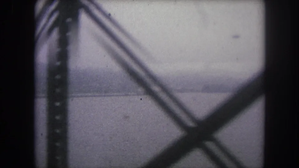 1962 8mm travel frame from Winter Park, Colorado, looking through dark rail bridge supports toward a gray river and hazy far bank.