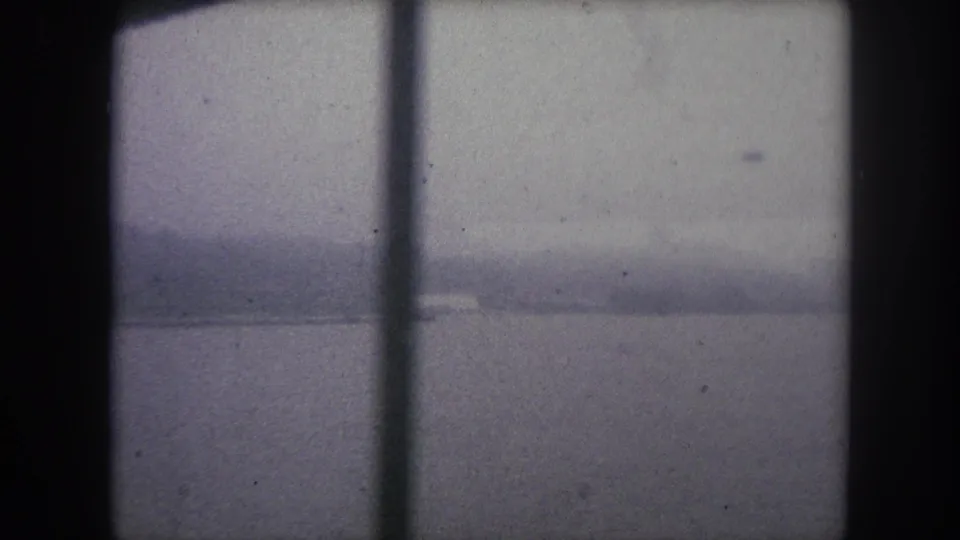 1962 8mm frame from a moving bridge view, with a vertical support, hazy river, low hills, and a faint light building on the far shore.