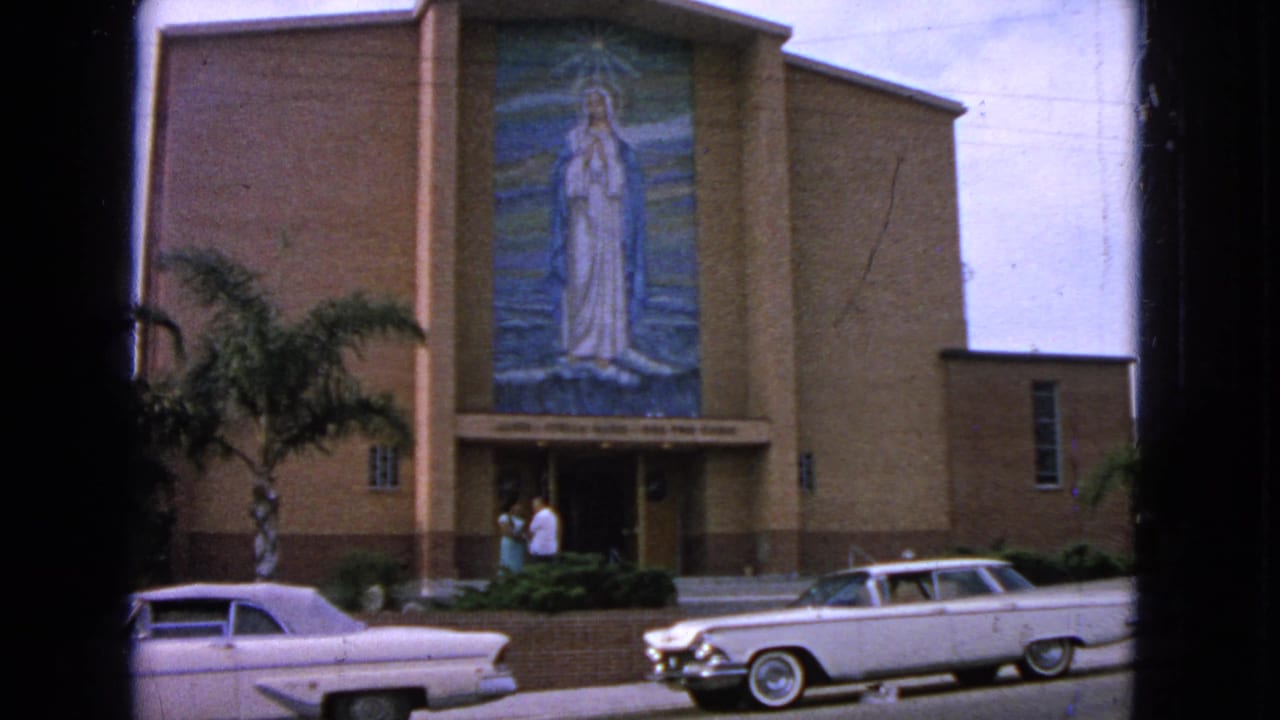 Brick church facade with Virgin Mary mosaic, two white cars, palm trees, and people near the entrance.
