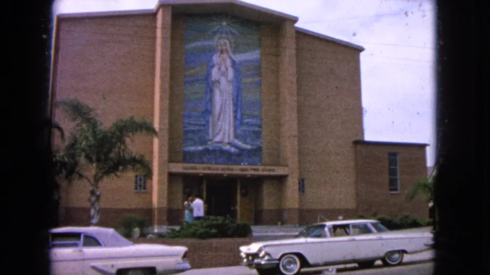 1963 home-movie frame of the Barrington church facade with Virgin Mary mosaic, palm tree, two adults near the doors, and white cars in front.