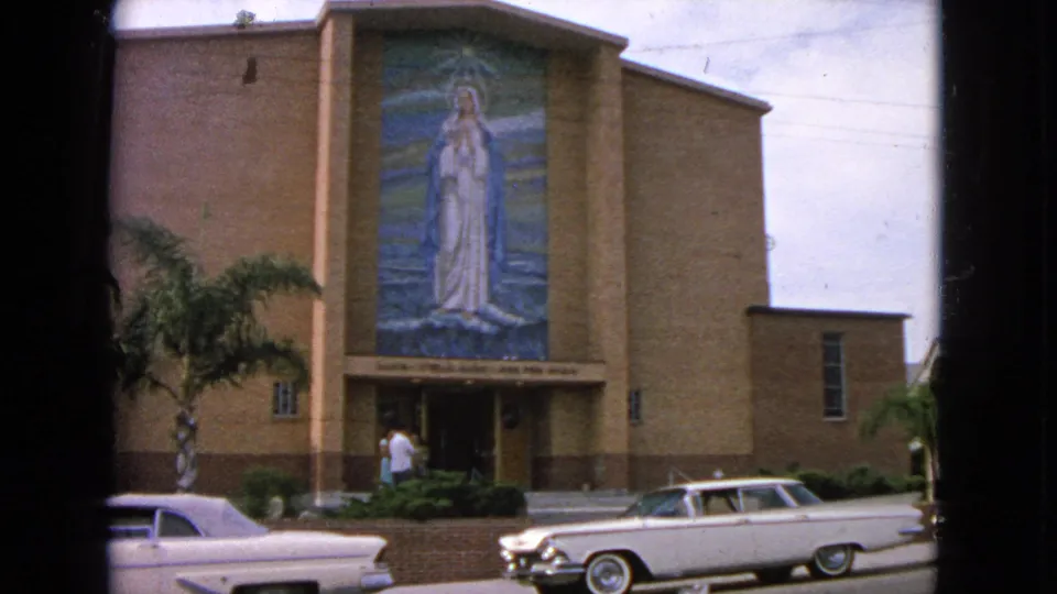 1963 home-movie view of a Barrington church exterior, showing the Marian mosaic, palm tree, front steps, and a white car near the curb.