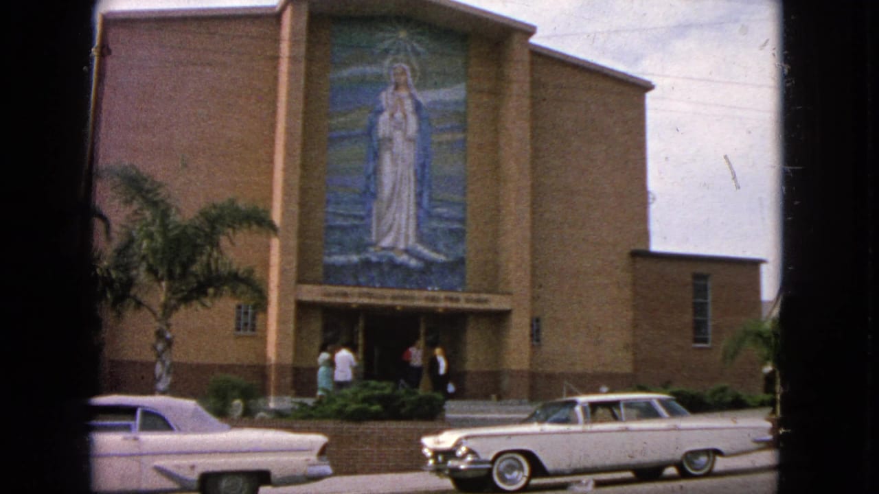 Church entrance with people outside, a Virgin Mary mural, palm trees, and classic cars in front.