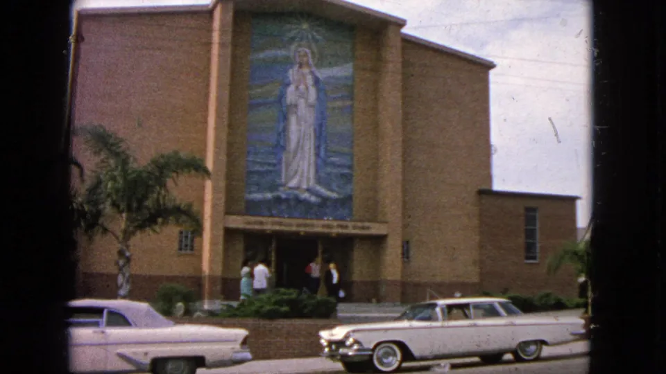Barrington church exterior in 1963 showing people near the entrance, a Virgin Mary mural above them, and early-1960s cars parked at the curb.