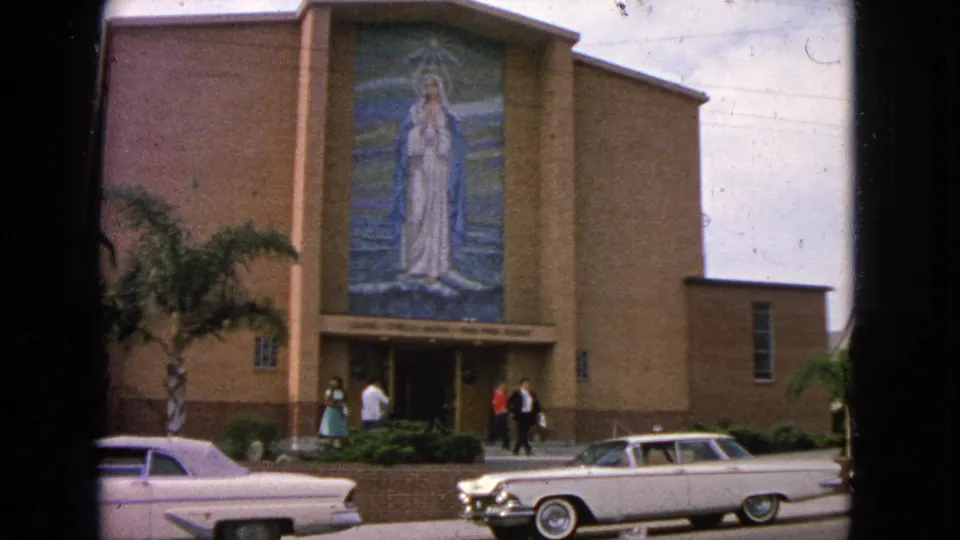 1963 home-movie frame of parishioners outside a Barrington church, with a large religious mural, brick walls, palm tree, and classic cars visible.